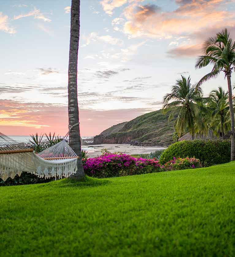 Atardecer desde un jardín del hotel las Alamandas, con una hamaca colocada desde en tronco de dos palmeras con vista al mar.
